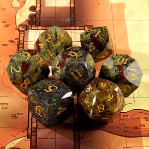 May include: A set of seven polyhedral dice with a green and red speckled pattern. The dice have gold-colored numbers and are arranged on a map-like surface. The dice are designed for tabletop role-playing games.