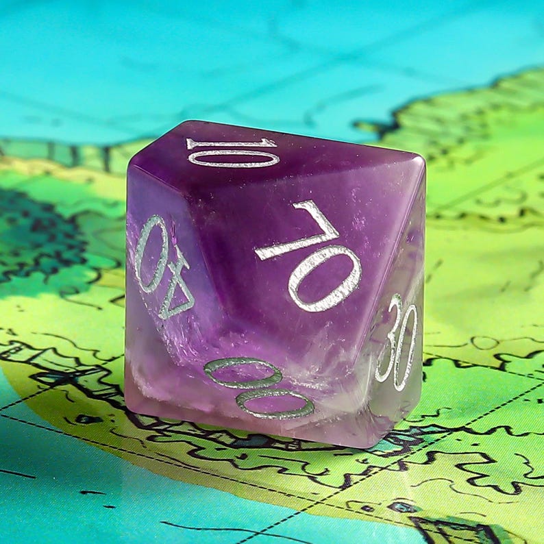 May include: A purple, 10-sided dice with white numbers. The dice is made of a translucent material, and the numbers are visible through the material.