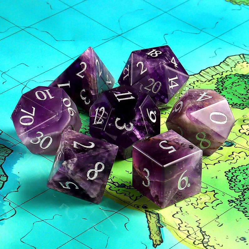 May include: Set of seven purple and white dice with various numbers on each side. The dice are made of amethyst and have a natural, textured surface.