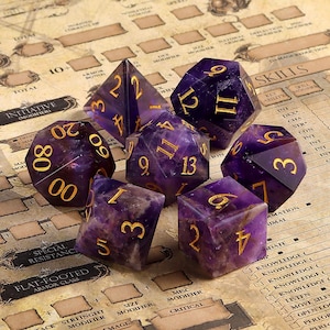 May include: Set of seven purple and gold dice with numbers 1-20. The dice are made of amethyst and have a natural, polished finish.