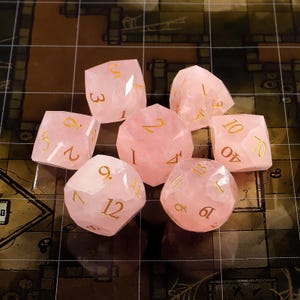 May include: A set of seven rose quartz dice with gold-colored numbers. The dice are in various geometric shapes, including a d4, d6, d8, d10, d12, d20, and a percentile die. The dice are arranged on a grid-patterned surface.