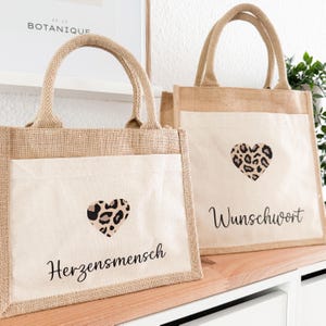 Könnte beinhalten: Zwei braune Jute-Tragetaschen mit weißen Stoffbahnen. Die Bahnen zeigen ein Herz im Leopardenmuster und schwarzen Text. Eine Tasche sagt "Herzensmensch" und die andere sagt "Wunschwort".