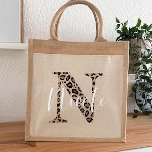 Könnte beinhalten: Eine natürliche Jute-Tragetasche mit einem weißen Canvas-Einsatz. Der Einsatz zeigt einen großen Buchstaben "N" in einem Leopardenmuster. Der Name "Nicole" ist in weißer Schrift unter dem Buchstaben geschrieben.