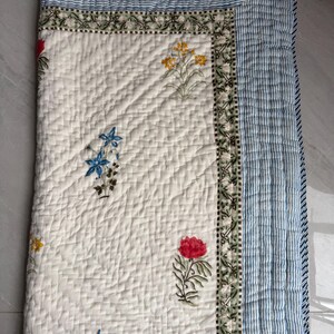 May include: A quilted, off-white blanket with a floral pattern. The blanket has a border of blue and white stripes and a decorative floral border. The floral pattern includes red, yellow, and blue flowers.