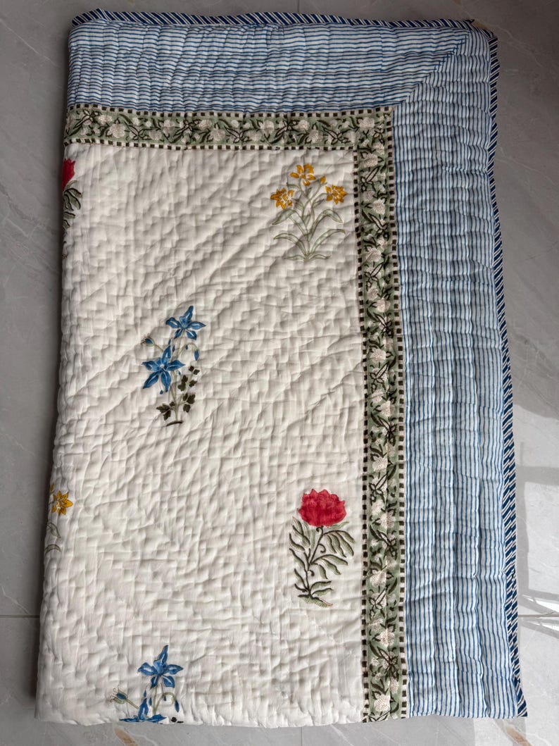 May include: A quilted, off-white blanket with a floral pattern. The blanket has a border of blue and white stripes and a decorative floral border. The floral pattern includes red, yellow, and blue flowers.