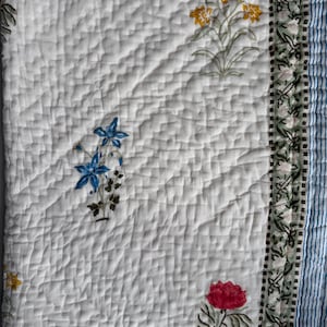 May include: White quilted textile with floral patterns in red, yellow, and blue. The quilt features a diamond pattern and a border with a green floral design. A blue and white striped edge is visible.