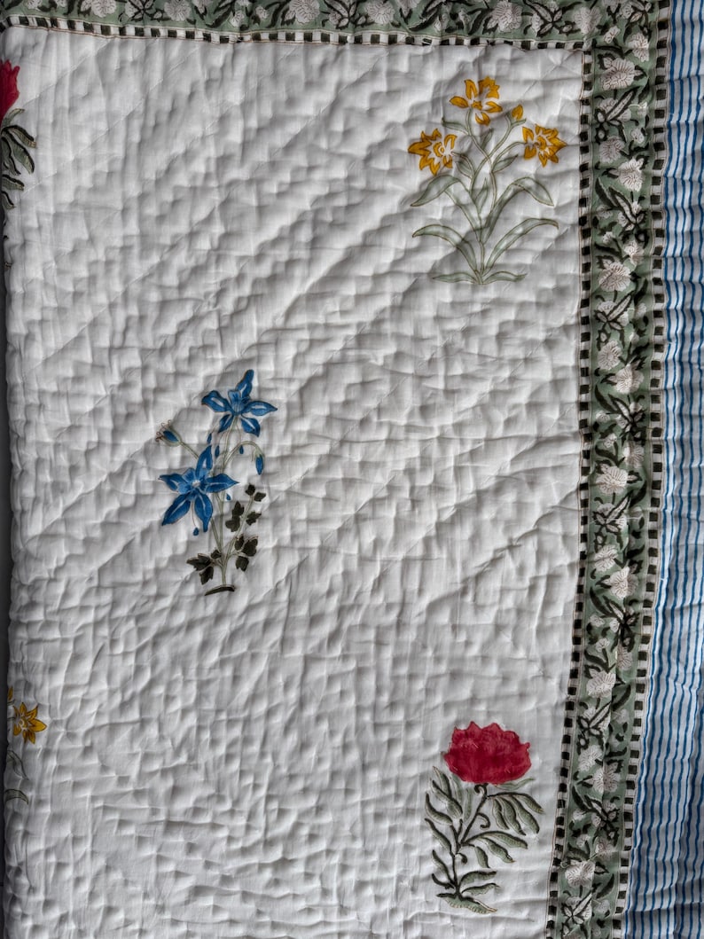 May include: White quilted textile with floral patterns in red, yellow, and blue. The quilt features a diamond pattern and a border with a green floral design. A blue and white striped edge is visible.