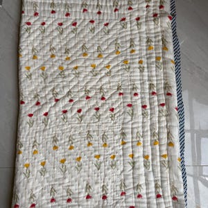 May include: A white quilted blanket featuring a floral design of red and yellow flowers. The blanket has a navy blue and white striped border. The flowers are embroidered on the blanket.