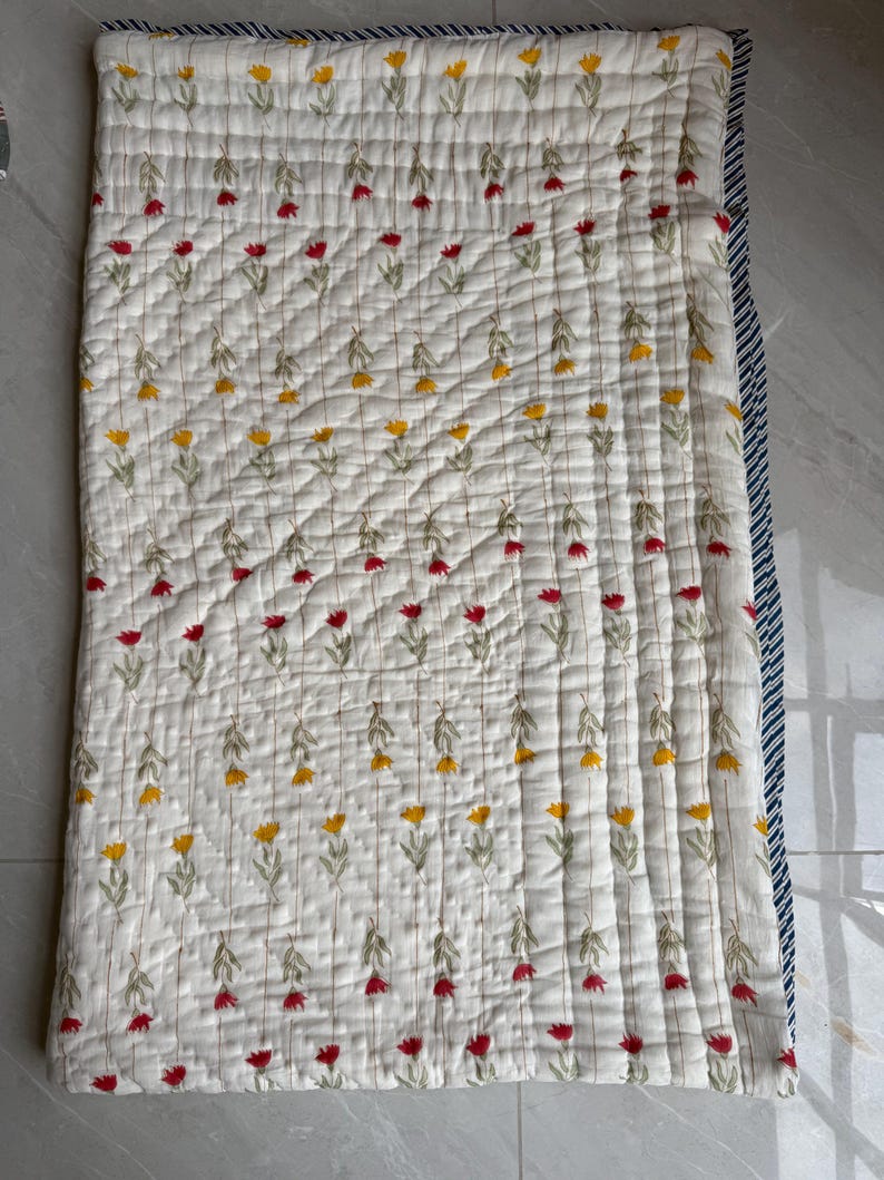 May include: A white quilted blanket featuring a floral design of red and yellow flowers. The blanket has a navy blue and white striped border. The flowers are embroidered on the blanket.
