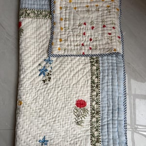 Może przedstawiać: Pikowana kołdra z kwiatowym wzorem. Kołdra ma białe tło z czerwonymi, niebieskimi i żółtymi kwiatowymi wzorami. Obramowanie ma niebiesko-białe paski. Kołdra wykonana jest z miękkiego, pikowanego materiału. Wymiary w centymetrach.