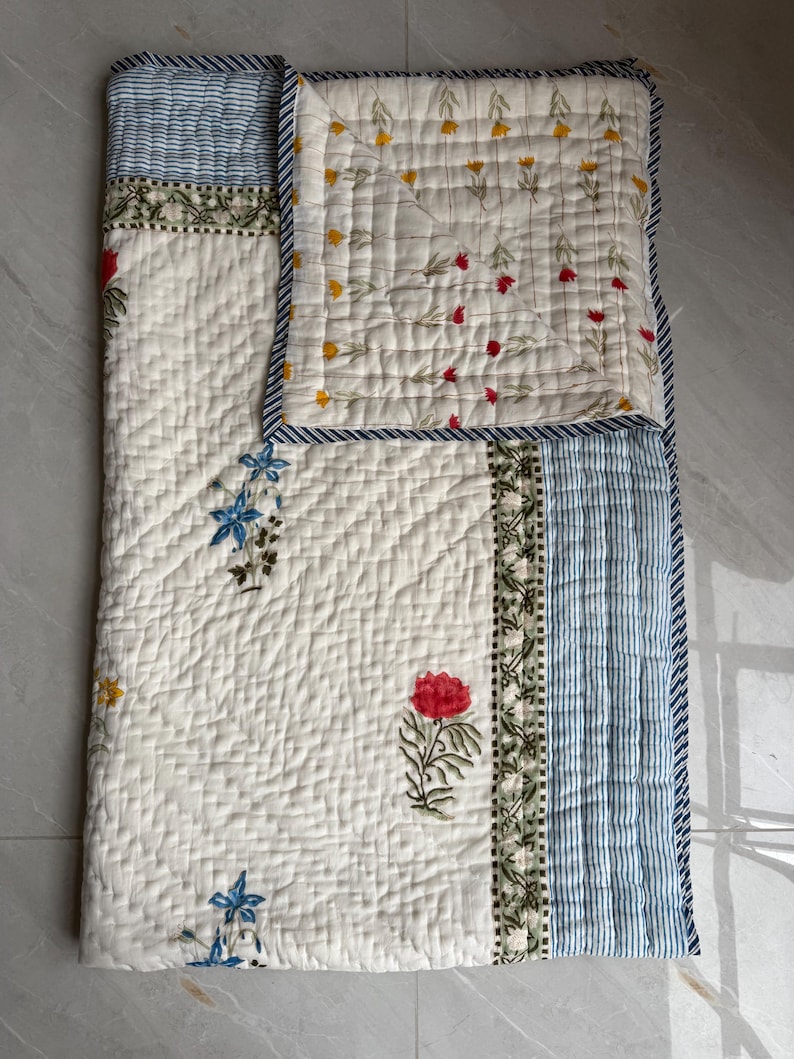 May include: A floral patterned quilt. The quilt has a white base with red, blue, and yellow flower designs. The border features blue and white stripes. The quilt is made of a soft, quilted material. The dimensions are in centimeters.