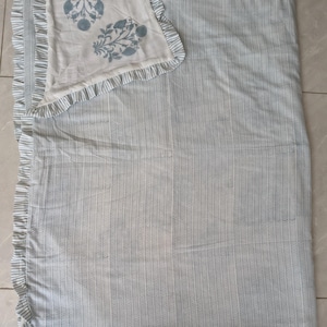 Peut inclure: Une couverture rectangulaire avec un motif à rayures chevron bleu clair et blanc et un bord à volants. Le coin supérieur est replié pour révéler un envers blanc orné de deux motifs floraux bleus imprimés sur la surface du tissu.