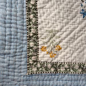 May include: A quilted textile with a white background and floral patterns in yellow, green, and blue. The border features a green floral design, and the edge has blue and white stripes. The quilt is likely for bedding or home decor.