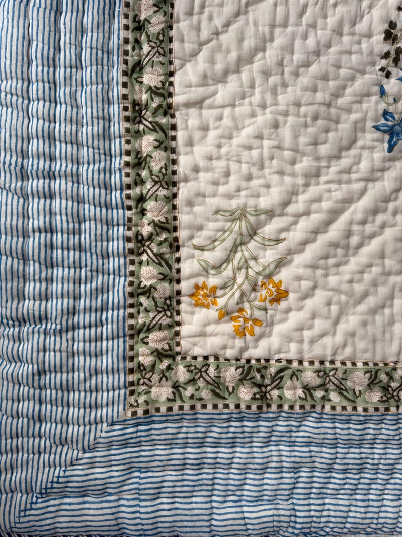 May include: A quilted textile with a white background and floral patterns in yellow, green, and blue. The border features a green floral design, and the edge has blue and white stripes. The quilt is likely for bedding or home decor.