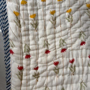 May include: A white quilted blanket with a floral pattern. The design features yellow and red flowers with green leaves, arranged on a striped background. The border is navy blue and white striped.