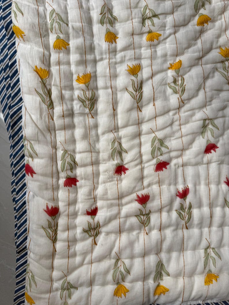 May include: A white quilted blanket with a floral pattern. The design features yellow and red flowers with green leaves, arranged on a striped background. The border is navy blue and white striped.