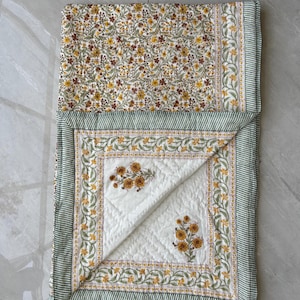 May include: A folded quilt with a floral pattern and a green and white striped border. The quilt features a white centre with yellow floral designs and a diamond-stitched pattern. The top section displays a dense floral print in yellow, brown, and green.