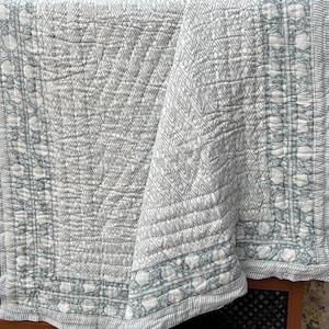 May include: A white and gray quilted blanket with a geometric diamond pattern. The border features a floral design and striped edges. The blanket is folded, showing its texture and design.