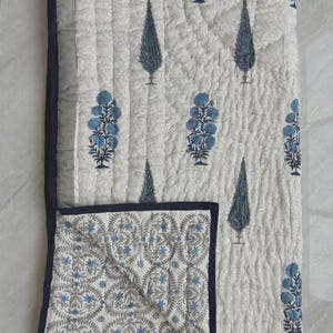 Peut inclure: Une couverture matelassée en coton avec une base blanche ornée d'un motif répétitif de fleurs bleues et de cyprès bleu foncé. La couverture présente une bordure bleu foncé et un coin replié révélant un motif secondaire de petites fleurs.