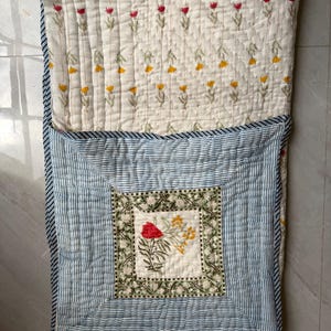 May include: A floral quilted blanket. The top section has a cream background with rows of red and yellow flower designs. The bottom section features blue and white stripes, with a central square displaying a floral motif. The blanket is made of soft fabric.