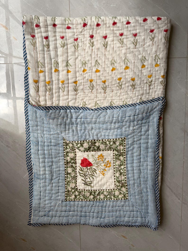 May include: A floral quilted blanket. The top section has a cream background with rows of red and yellow flower designs. The bottom section features blue and white stripes, with a central square displaying a floral motif. The blanket is made of soft fabric.