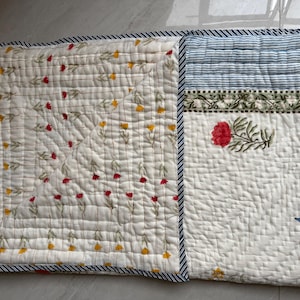 May include: A quilted textile in off-white, adorned with floral and striped patterns. The left section displays a repeating pattern of red and yellow flowers. The right side features a red floral design, blue stripes, and a blue floral accent. The edges are trimmed with a blue and white striped border.