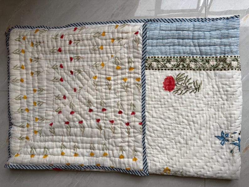 May include: A quilted textile in off-white, adorned with floral and striped patterns. The left section displays a repeating pattern of red and yellow flowers. The right side features a red floral design, blue stripes, and a blue floral accent. The edges are trimmed with a blue and white striped border.