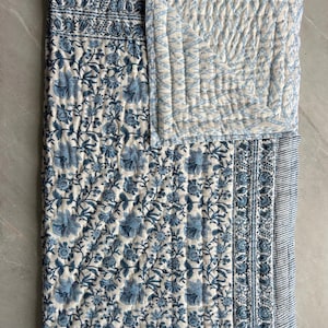 May include: A blue and white floral patterned quilt. The quilt features a repeating pattern of blue flowers and leaves on a white background. The border has a striped design. The quilt is folded over, revealing a quilted section with a geometric pattern.