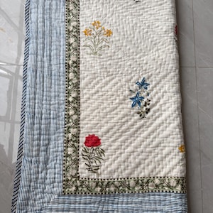May include: A white quilted blanket with floral embroidery in red, yellow, and blue. The blanket has a blue and white striped border and a decorative floral border. The quilt is folded and appears to be made of soft fabric.