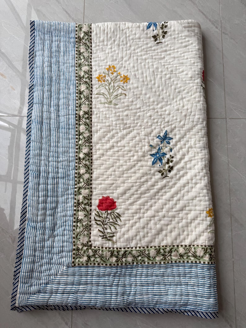 May include: A white quilted blanket with floral embroidery in red, yellow, and blue. The blanket has a blue and white striped border and a decorative floral border. The quilt is folded and appears to be made of soft fabric.