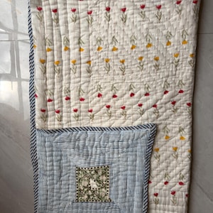 May include: Cream quilt with a floral pattern of red and yellow flowers. A blue and white striped section features a square floral design. The edges are trimmed with a blue and white striped border. The quilt is made of soft fabric.