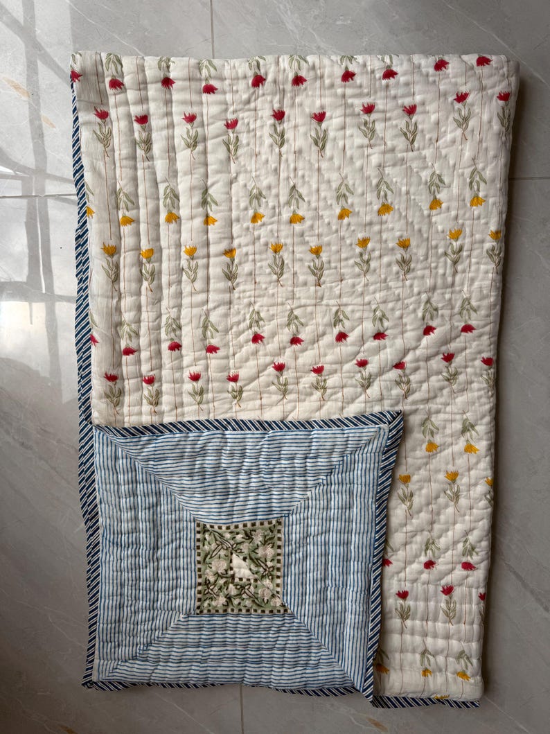 May include: Cream quilt with a floral pattern of red and yellow flowers. A blue and white striped section features a square floral design. The edges are trimmed with a blue and white striped border. The quilt is made of soft fabric.