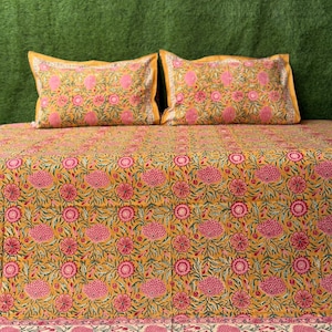 Puede incluir: Una sábana de cama de color amarillo mostaza con un estampado floral rosa. La sábana tiene un borde con un estampado floral más pequeño en rosa, blanco y verde. También se muestran dos fundas de almohada a juego.