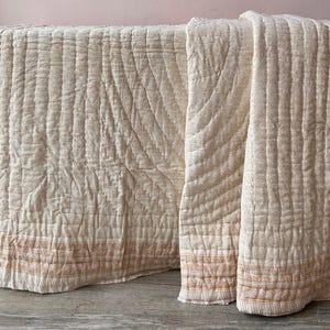 May include: A cream-colored quilted throw blanket with a subtle orange and white striped border. The blanket is folded and displayed on a light brown surface.