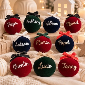 May include: A collection of ten spherical velvet Christmas ornaments in red, dark green, and navy blue. Each ornament features a personalized name in white or gold script, topped with a small decorative ribbon bow and a gold metal hanging loop.