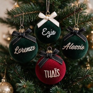 May include: Four spherical Christmas ornaments with a velvet texture. Three are dark green with black bows and gold lettering reading Lorenzo, Ezio, and Alessio. One is deep red with a black bow and gold lettering reading THA&Iuml;S.