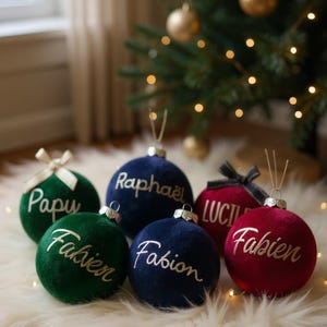 May include: A collection of six spherical velvet Christmas ornaments in deep green, navy blue, and burgundy red. Each ornament features a metallic cap with a hanging loop and is personalized with white or gold cursive text including names like Papu, Rapha&euml;l, Fabien, and Lucie.