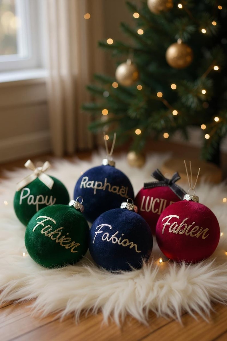 May include: A collection of six spherical velvet Christmas ornaments in deep green, navy blue, and burgundy red. Each ornament features a metallic cap with a hanging loop and is personalized with white or gold cursive text including names like Papu, Rapha&euml;l, Fabien, and Lucie.