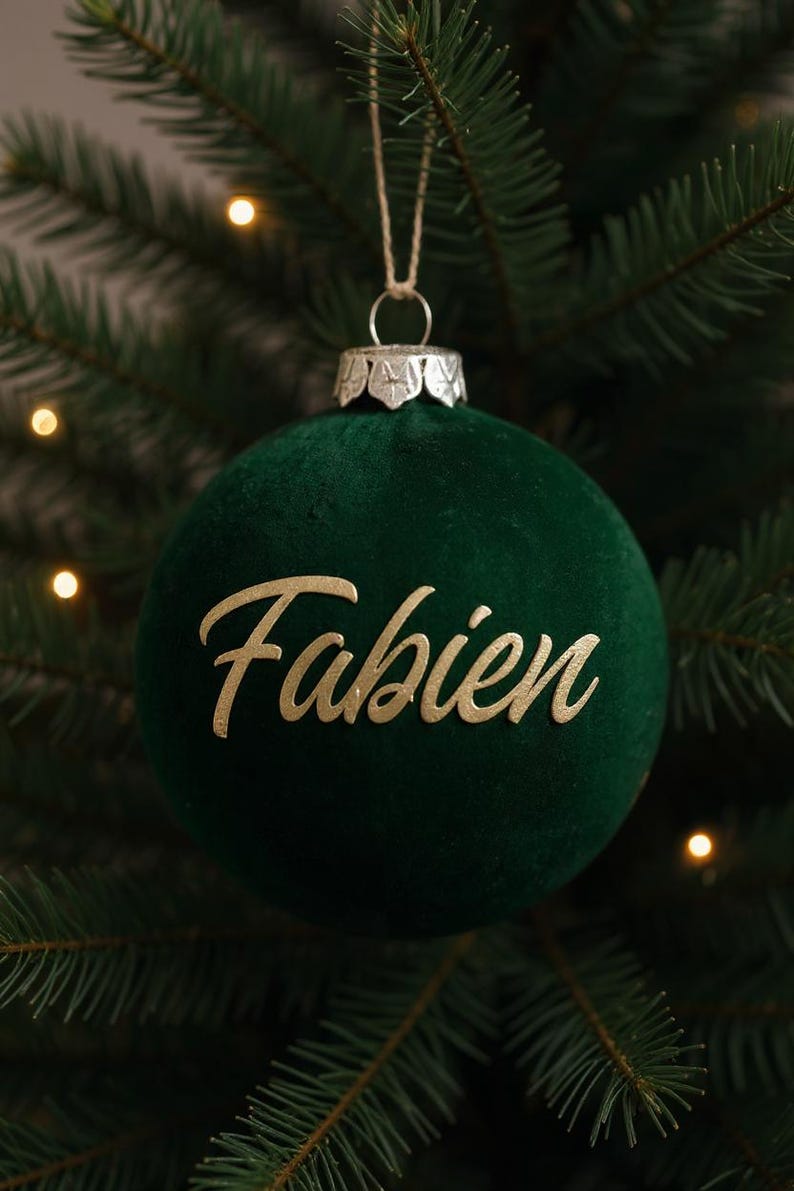 May include: A spherical Christmas ornament covered in dark green velvet fabric. The name Fabien is written across the center in gold-colored cursive lettering. A silver-toned metal cap sits at the top with a thin string attached for hanging.