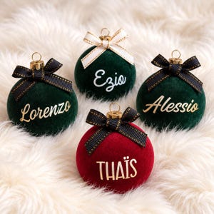 May include: Four spherical ornaments with a velvet texture. Three are dark green with gold or white lettering reading Lorenzo, Ezio, and Alessio. One is red with gold lettering reading THA&Iuml;S. Each features a gold cap and a decorative ribbon bow.