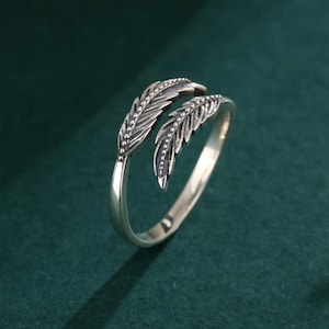 Puede incluir: Un anillo plateado con diseño de pluma. El anillo presenta dos detalles de plumas estilizadas con texturas detalladas. El anillo está sobre un fondo verde oscuro, con el nombre de la marca "Labellry" visible en la esquina superior izquierda.