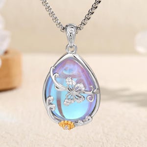 May include: Silver-toned necklace with a teardrop pendant. The pendant features a pearlescent, iridescent stone with a butterfly design and floral accents. The bottom of the pendant has a small gold-colored accent. The necklace is displayed on a light-colored surface.