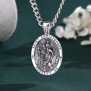 Könnte beinhalten: Eine silberfarbene ovale Anhängerkette mit einer geprägten Darstellung des heiligen Christophorus, der ein Kind trägt. Der Anhänger ist mit dem Text SAINT CHRISTOPHER PROTECT US versehen und hängt an einer passenden silberfarbenen Gliederkette.