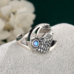 Puede incluir: Un anillo de plata con un diseño de lobo aullando sobre una luna creciente. El anillo tiene una cabeza de lobo detallada y una pequeña gema azul redonda. El anillo se muestra sobre una superficie gris suave.