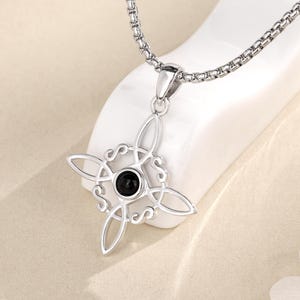 以下が含まれることがあります： 四角いデザインと透かし模様が特徴のシルバーペンダントネックレス。ペンダントの中央には、小さな丸い黒い石がセットされています。チェーンはリンクスタイルです。「Labellry Handmade Jewelry」のブランド名が表示されています。