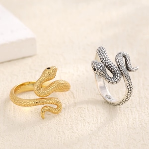 May include: Two snake-shaped rings. One is gold-toned, the other is silver-toned. Both rings feature a coiled snake design with textured scales and small black eyes. The rings are displayed on a light-coloured surface.