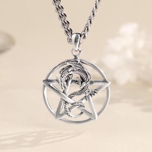 Può includere: Una collana con pendente argentato con un design a drago all'interno di un pentagramma. Il drago è finemente dettagliato e la collana ha una catena con un design a torsione. Il marchio "Labellry Handmade Jewelry" è visibile in alto a sinistra.