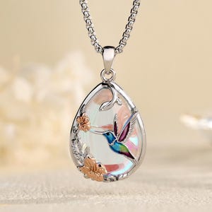 Puede incluir: Un collar de tono plateado con un colgante en forma de lágrima con un centro iridiscente. El colgante muestra un colibrí colorido y flores en tono oro rosa. El texto Labellry Handmade Jewelry es visible en la esquina superior derecha.