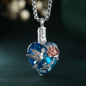 Peut inclure: Un collier en chaîne argent avec un pendentif en forme de cœur en cristal bleu. Le cœur présente un colibri en vol, une rose et de délicates vignes argentées.