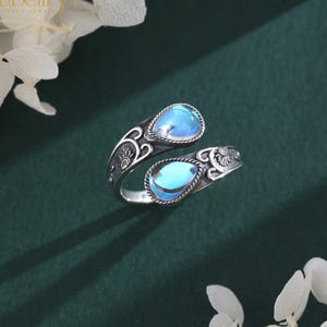 May include: An open-style silver ring featuring two teardrop-shaped iridescent blue stones. The metal band displays intricate oxidized floral engravings and scrollwork details, creating a textured appearance around the two prominent blue gems.